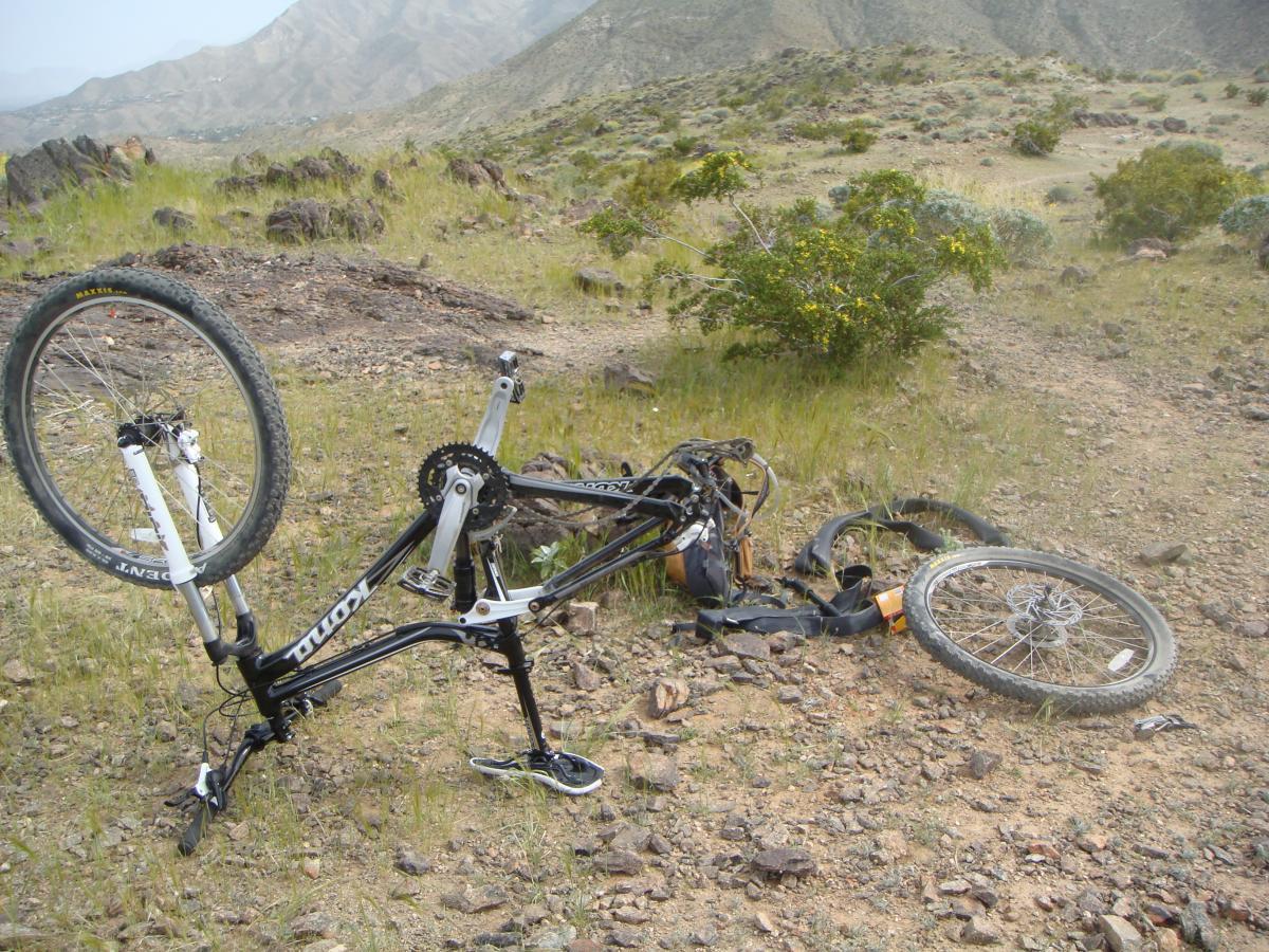 A fallen mountain bike lies on rocky terrain, partially obscured by grass and small bushes, with a mountainous landscape in the background. One wheel is upright, while the frame is on its side, suggesting the bike has been recently abandoned or crashed. The Goat Trails mountain bike trail.