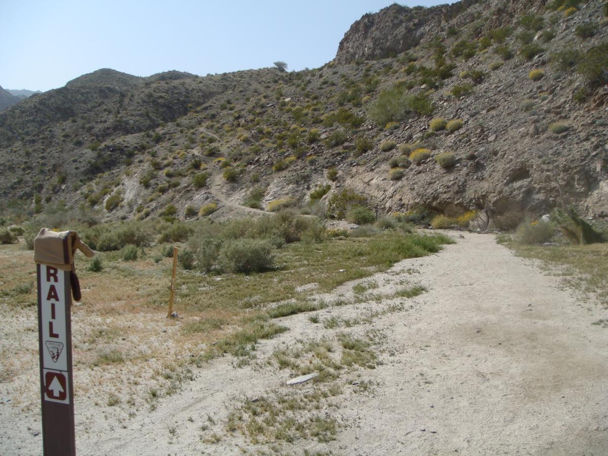 A dirt pathway leading into a hilly desert landscape, with sparse vegetation and rocky terrain. A wooden signpost marked "RAIL" stands in the foreground, indicating the direction of the trail. The atmosphere is sunny and clear, showcasing the rugged natural beauty of the area. Art Smith Trail mountain bike trail.