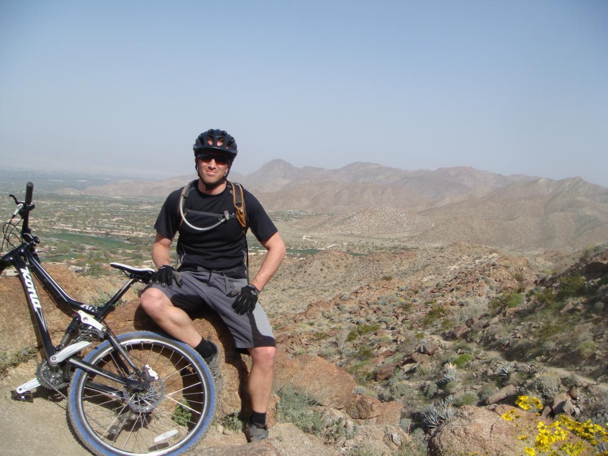 A mountain biker wearing a helmet and gloves sits on a rock, posing next to a black mountain bike on a rocky trail. The background features a panoramic view of hills and valleys under a clear sky. Art Smith Trail mountain bike trail.
