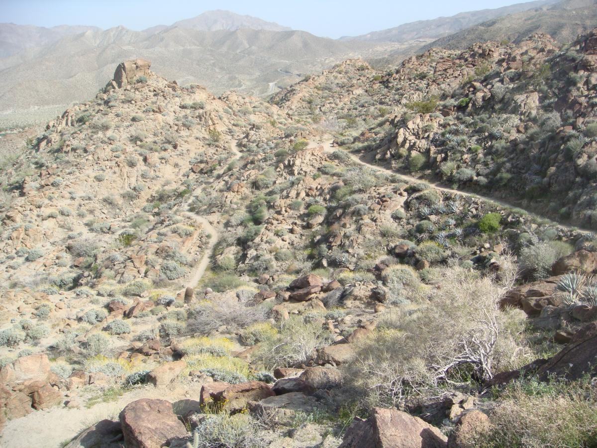 A scenic view of rugged mountain terrain featuring rocky landscapes, sparse vegetation, and winding dirt trails. The background showcases rolling hills and distant mountains under a clear sky. Art Smith Trail mountain bike trail.