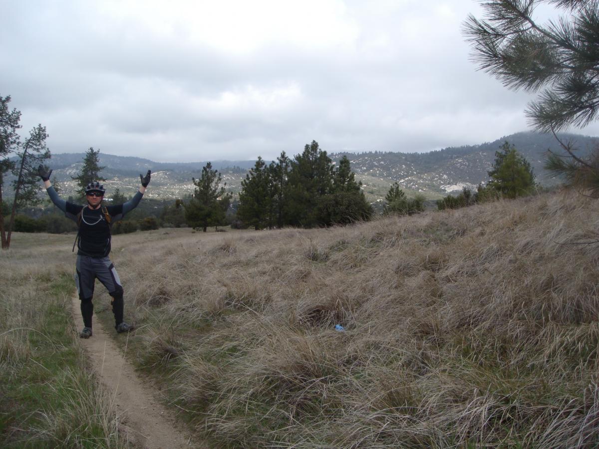 A person wearing a bike helmet, sunglasses, and cycling gear stands on a dirt path in a grassy area, raising their arms in celebration. Trees and distant hills are visible in the background under a cloudy sky. Exfoliator mountain bike trail.