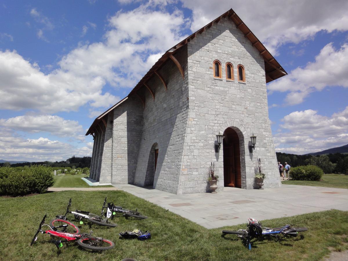 A stone building with a peaked roof stands against a blue sky with fluffy clouds. In the foreground, several bicycles lie on the grass, with a landscaped area featuring shrubs and a pathway leading up to the entrance of the building. People can be seen in the background enjoying the outdoor space. Kingdom Trails mountain bike trail.