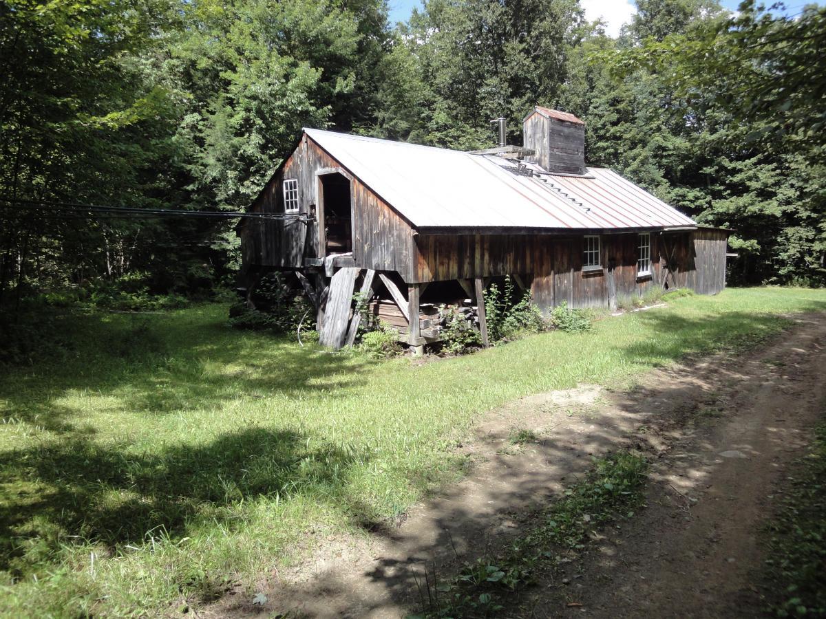 Alt text: A rustic wooden building with a metal roof, surrounded by lush green trees and grass. A dirt path leads up to the structure, which has a single open door and a chimney on the roof. Kingdom Trails mountain bike trail.
