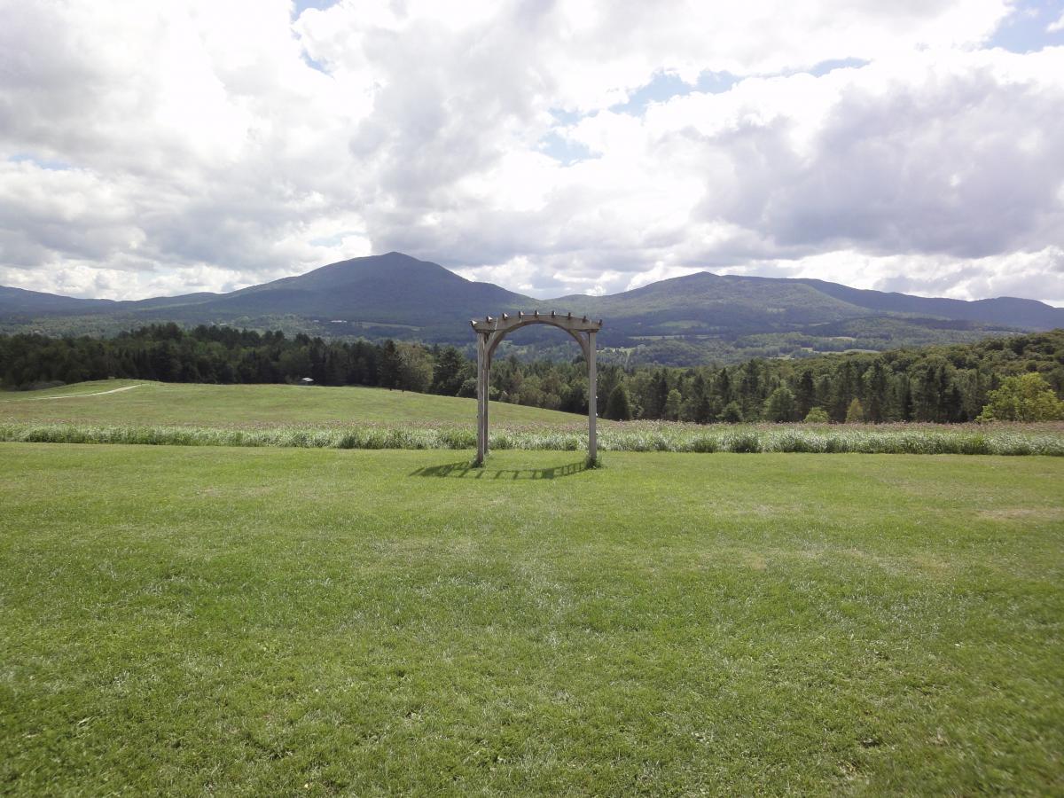 A wooden arbor stands in a lush green field, with rolling hills and mountains in the background under a partly cloudy sky. The landscape features a vibrant expanse of grass and trees, creating a serene natural setting. Kingdom Trails mountain bike trail.