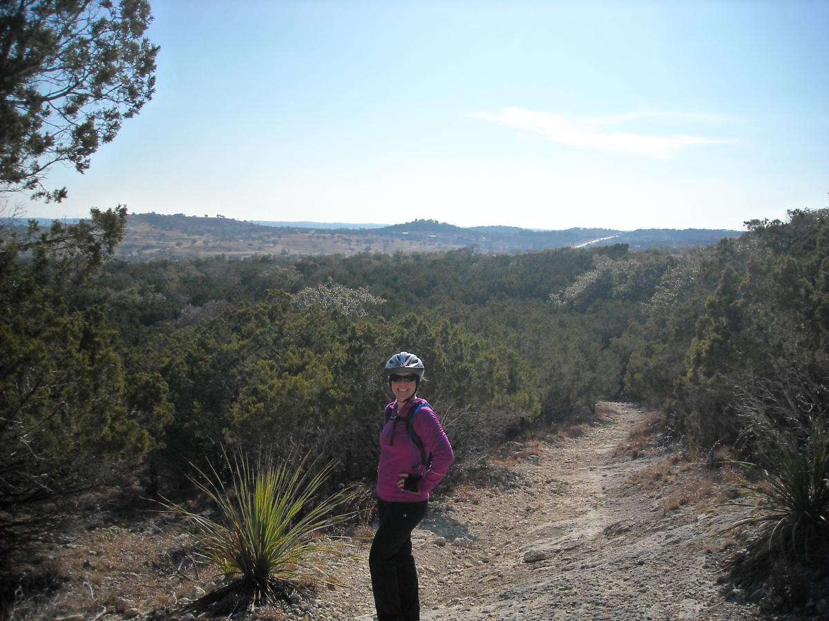 A person wearing a helmet and a pink jacket stands on a dirt trail surrounded by green shrubs and trees, with a scenic landscape in the background under a clear blue sky. Kerrville Schreiner Park mountain bike trail.