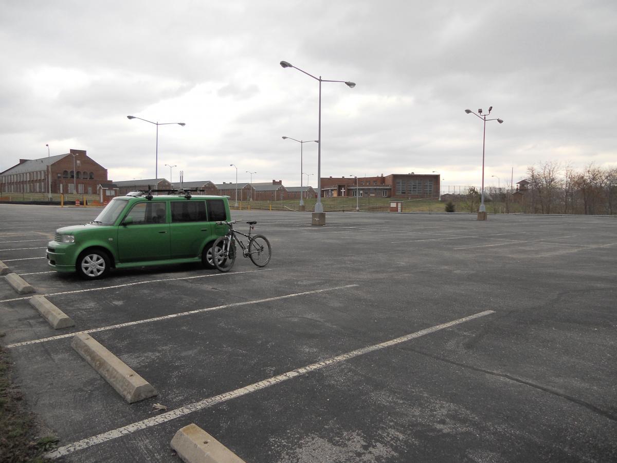 A green car parked in an empty lot with a bicycle attached to its side, under overcast skies. In the background, there are several brick buildings and parking lot lights, indicating a desolate urban environment. Laurel Hill Park mountain bike trail.