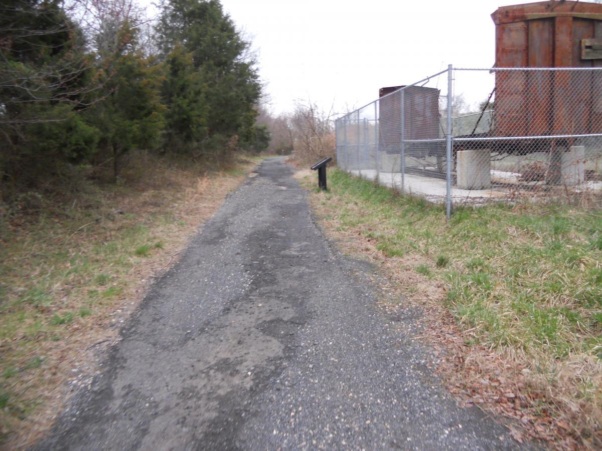 A gravel path bordered by overgrown grass and trees, leading towards a fenced area with old, rusted structures. A small informational sign is visible alongside the path. The scene is set under a gray, overcast sky. Laurel Hill Park mountain bike trail.