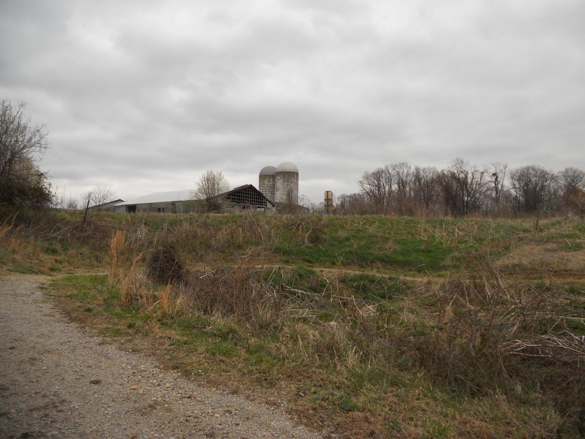 A rural landscape featuring a gravel path leading through tall, dry grasses. In the background, a weathered barn and two silos are visible under a cloudy sky, surrounded by bare trees and an open field. Laurel Hill Park mountain bike trail.