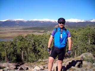 A person in cycling gear stands on rocky terrain with a scenic mountain range in the background. The mountains are capped with snow, and the landscape includes green trees and open fields under a clear blue sky. West Jefferson Trail mountain bike trail.