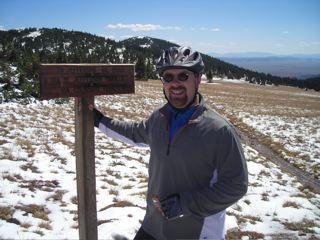 A man wearing a bike helmet and sunglasses stands next to a wooden sign on a snowy hillside, with mountains in the background. He is smiling and dressed in a gray jacket. The landscape features grassy areas and distant hills under a clear blue sky. West Jefferson Trail mountain bike trail.