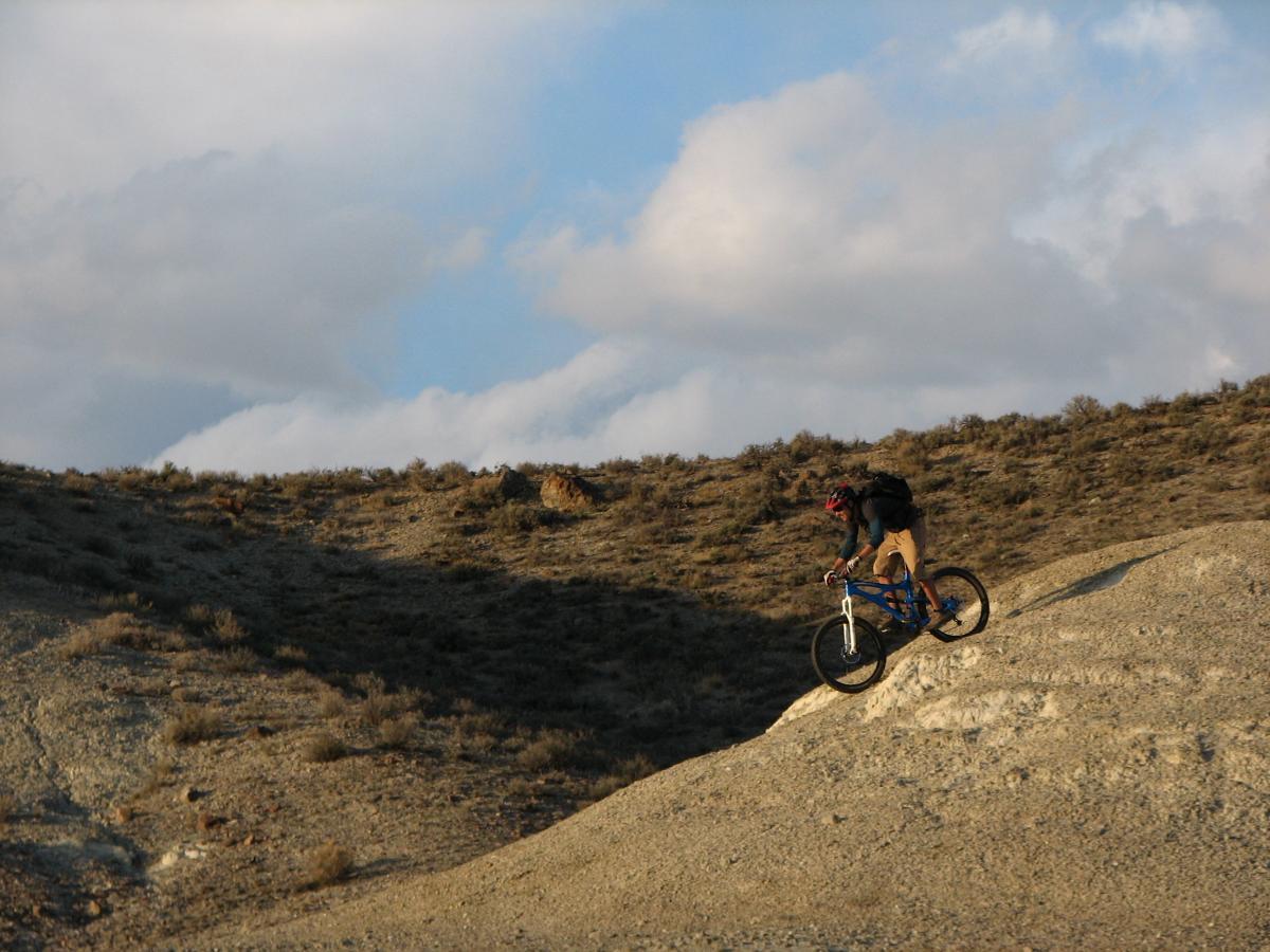 A mountain biker riding down a rocky slope in a semi-desert landscape under a partly cloudy sky. The cyclist is wearing a helmet and has a backpack, showcasing an adventurous outdoor activity. Discovery Hill Trails mountain bike trail.