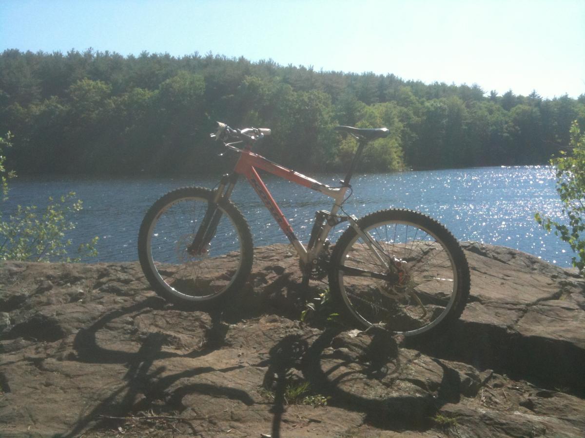 A mountain bike resting on a rocky shoreline beside a shimmering blue lake, with green trees and a clear sky in the background. Middlesex Fells Reservation mountain bike trail.