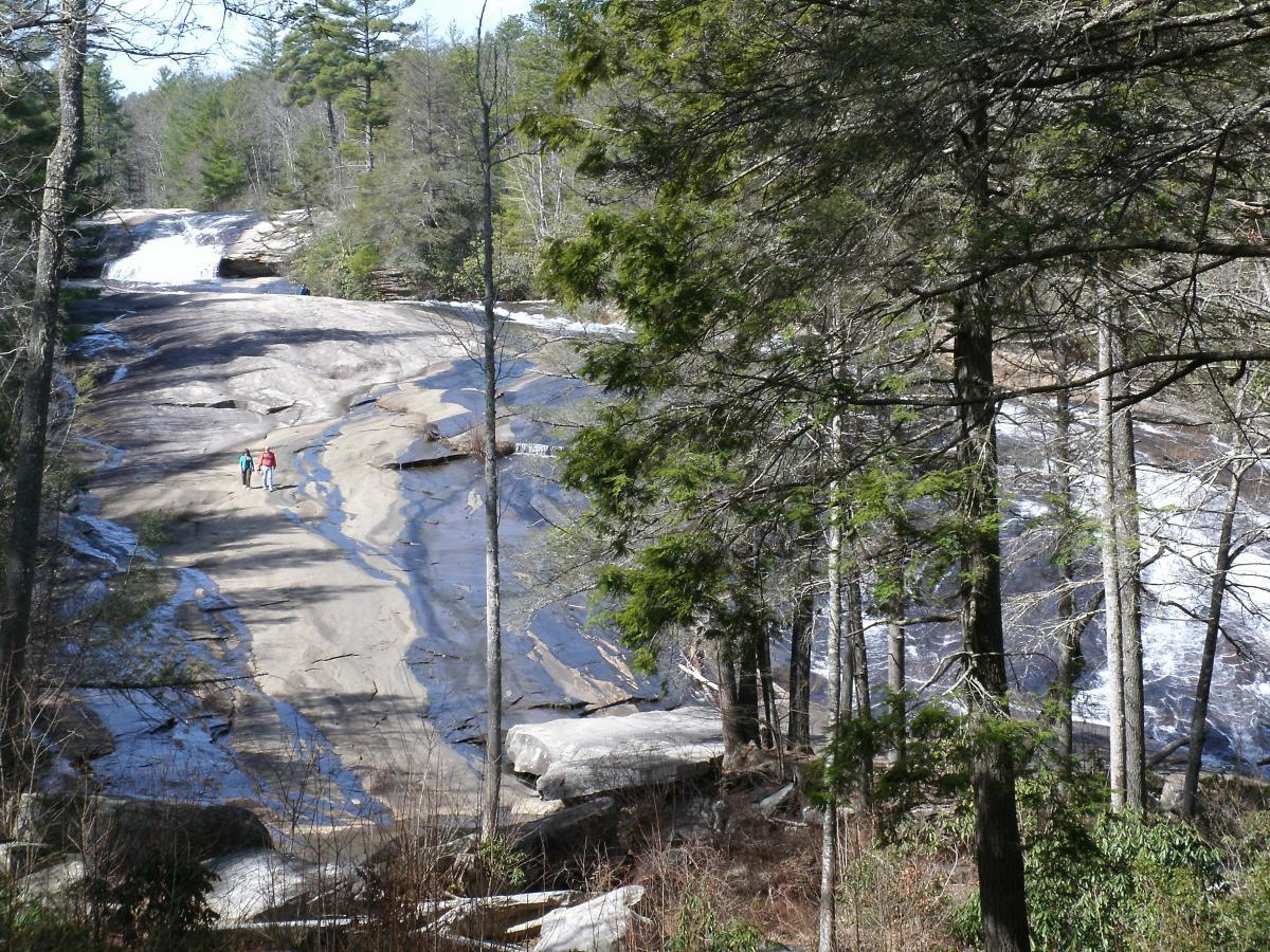 A scenic view of a rocky landscape featuring a small waterfall in the background. Two people walk along a smooth, wet rock surface, with lush green trees and a forested area surrounding them. The scene captures the beauty of nature in a tranquil outdoor setting. DuPont State Forest mountain bike trail.