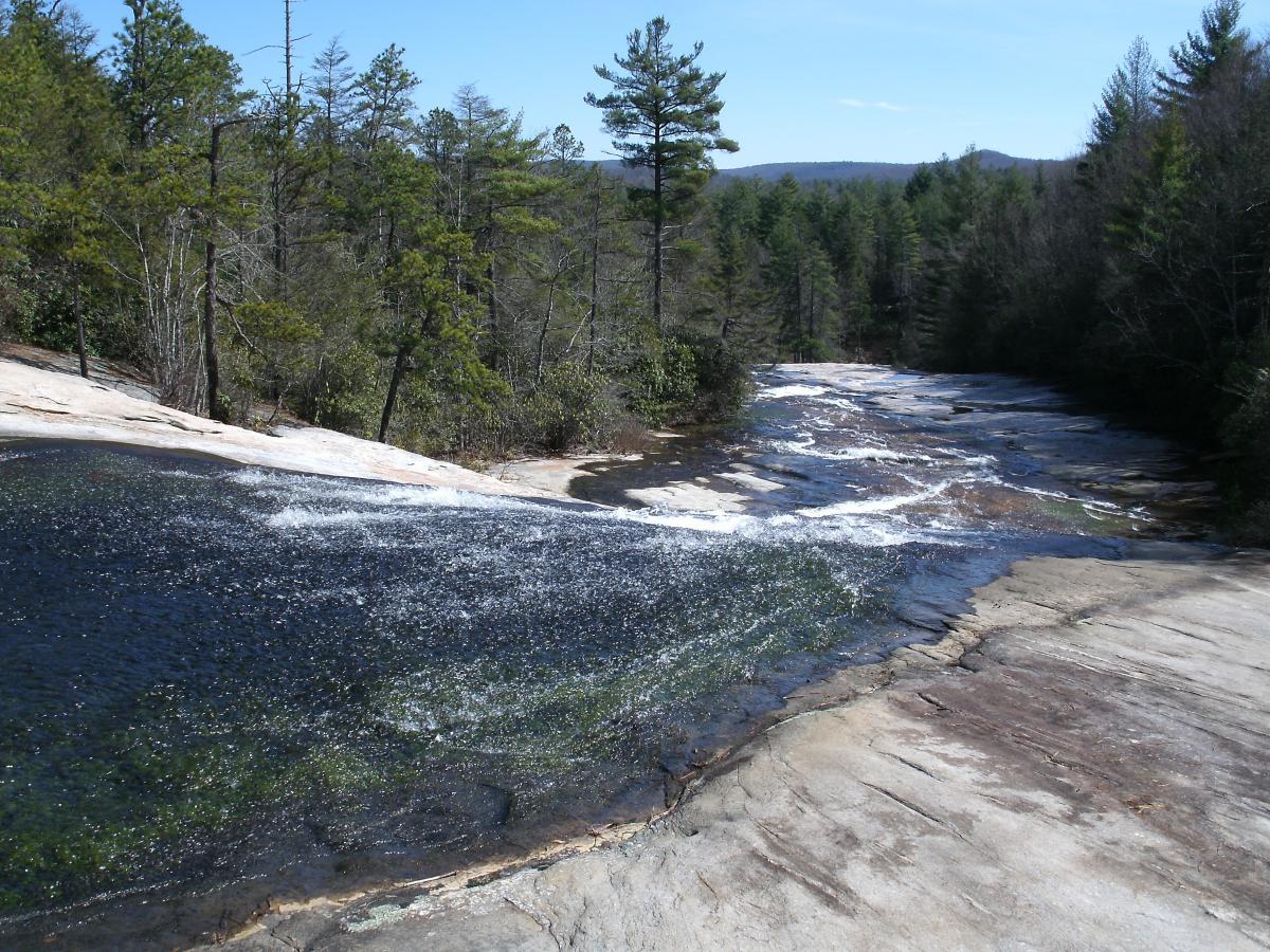 A serene landscape featuring a smooth, sloped rock surface with a gentle waterfall cascading over its edge. The water flows into a clear pool, surrounded by lush green trees and a backdrop of distant mountains under a clear blue sky. DuPont State Forest mountain bike trail.