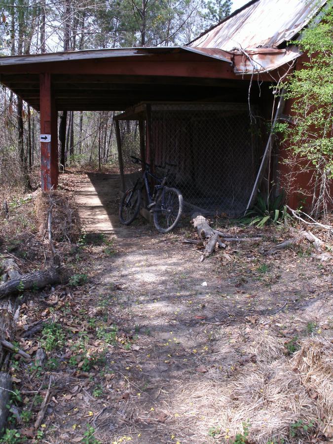 A dirt path leads to a covered area with a red wooden structure, partially covered by a corrugated metal roof. A black bicycle rests against one side of the structure, surrounded by scattered leaves and fallen branches, with trees in the background. Arrowhead Park mountain bike trail.