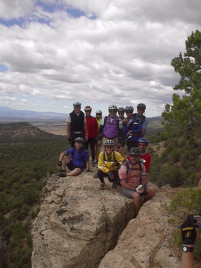 A group of cyclists wearing helmets and colorful jerseys poses on a rocky cliff overlooking a landscape of trees and valleys under a cloudy sky. Paradox Trail mountain bike trail.