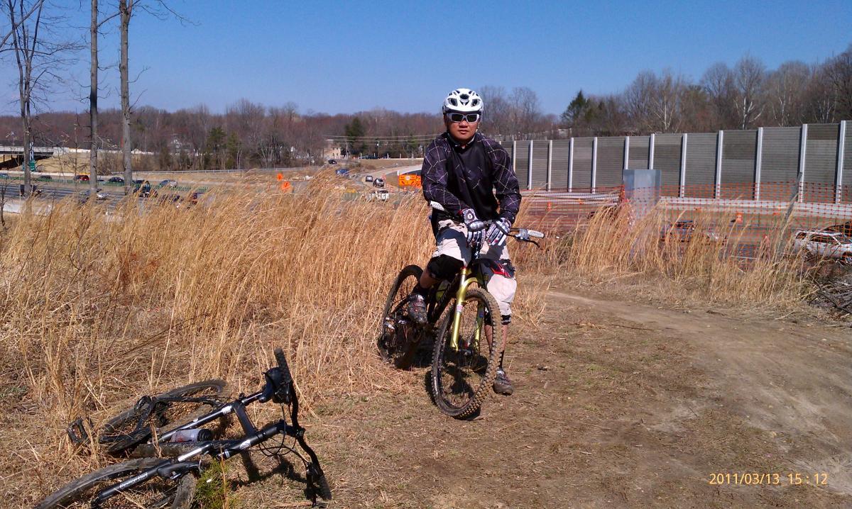 A person wearing a helmet and biking gear sits on a yellow mountain bike on a dirt trail, surrounded by tall grass. In the background, there are trees and a partially visible roadway with construction barriers and vehicles. The sky is clear and blue, indicating a sunny day. A second bike lies on the ground nearby. Wakefield mountain bike trail.