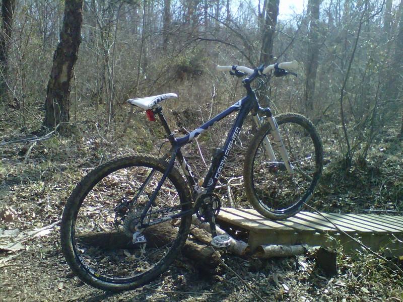 A mountain bike parked on a wooden bridge in a wooded area, surrounded by bare trees and earthy foliage. The bike shows signs of use, with mud on its tires. Sunlight filters through the trees, creating a serene outdoor setting. Rappahanock River Trail mountain bike trail.