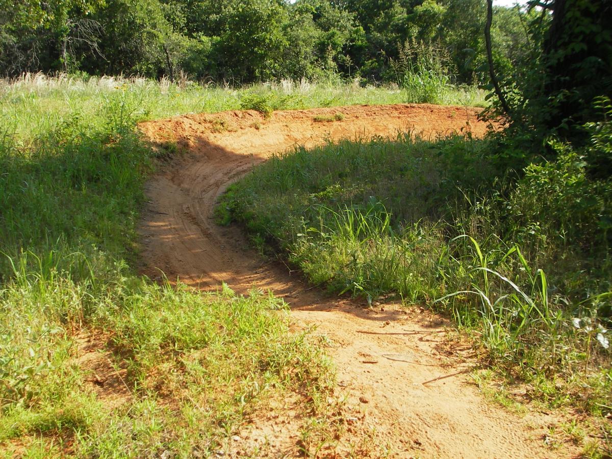 A sandy path curves through a grassy area, surrounded by lush greenery and trees in the background, indicating a natural outdoor setting. Lake Stanley Draper mountain bike trail.