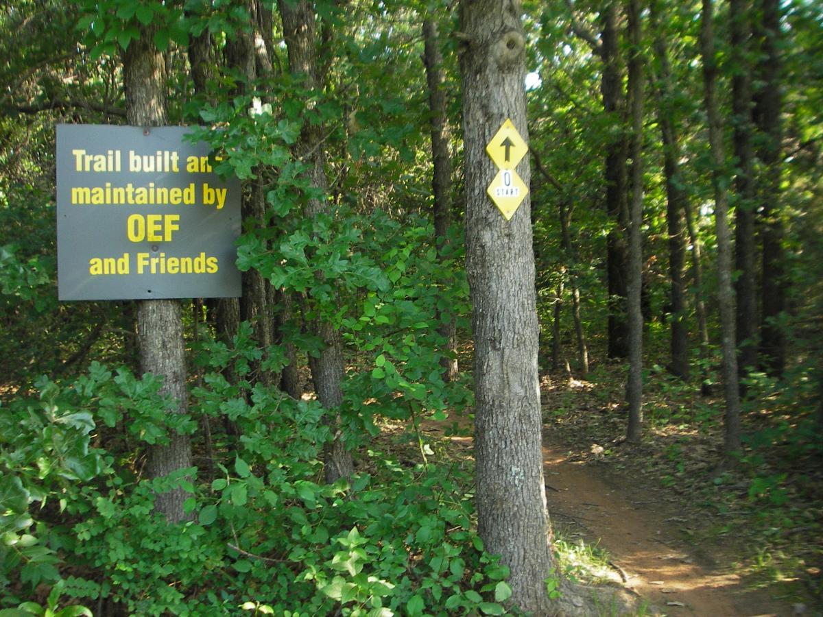 A sign indicating a trail built and maintained by OEF and Friends is situated among green foliage. Next to the sign, a yellow trail marker signifies the start of the path, which winds into a wooded area. Lake Stanley Draper mountain bike trail.