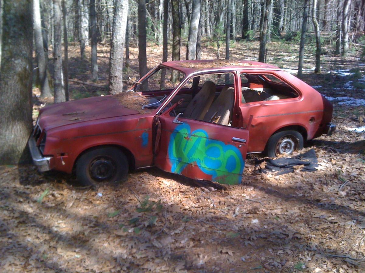 A rusted red car parked in a forested area, partially hidden by trees. The car has visible signs of wear, including graffiti on the side and leaves piled on top. The driver's side door is open, and one tire is flat, with debris scattered around on the ground. Willowdale Forest mountain bike trail.