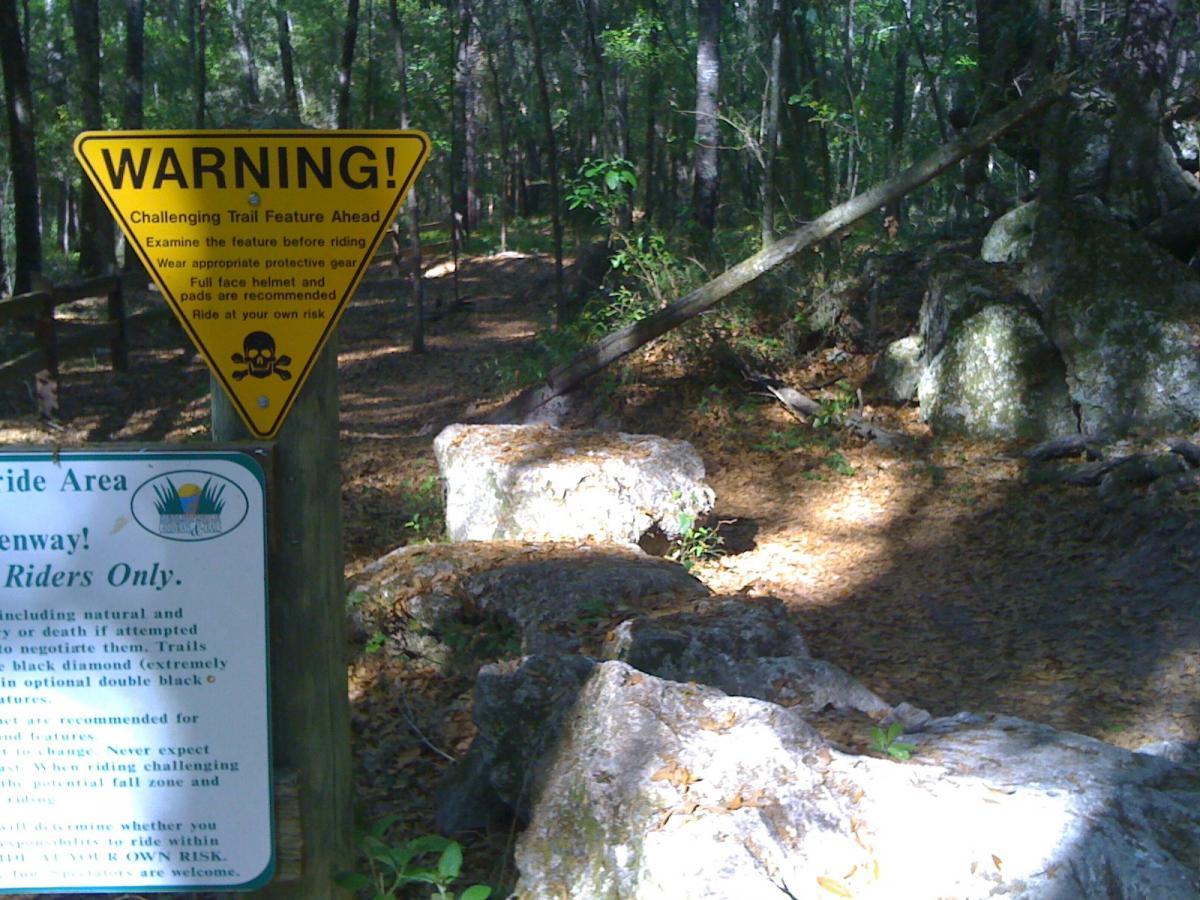 A triangular yellow warning sign indicating a challenging trail feature ahead, with advice to examine the feature before riding, wear protective gear, and ride at one's own risk. The background features a wooded trail with rocks and trees, and an additional sign promoting rider safety and guidelines. Santos mountain bike trail.