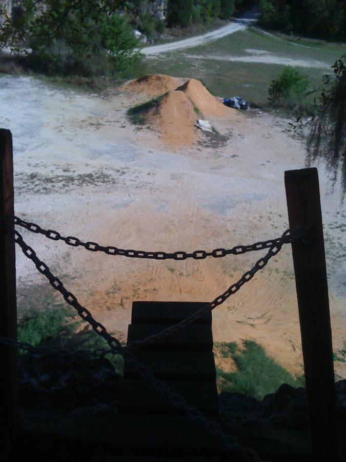 A view from a wooden platform, looking down at a dirt area with two small mounds of dirt, surrounded by grass and trees. A chain barrier is visible in the foreground, and a pathway can be seen in the background. Santos mountain bike trail.