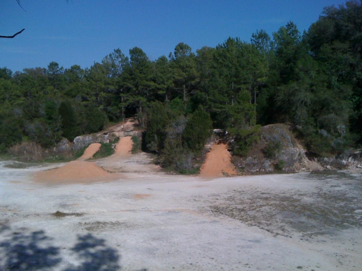 A landscape featuring a sandy area with reddish-brown soil slopes leading up to a forested backdrop of various green trees under a clear blue sky. Santos mountain bike trail.
