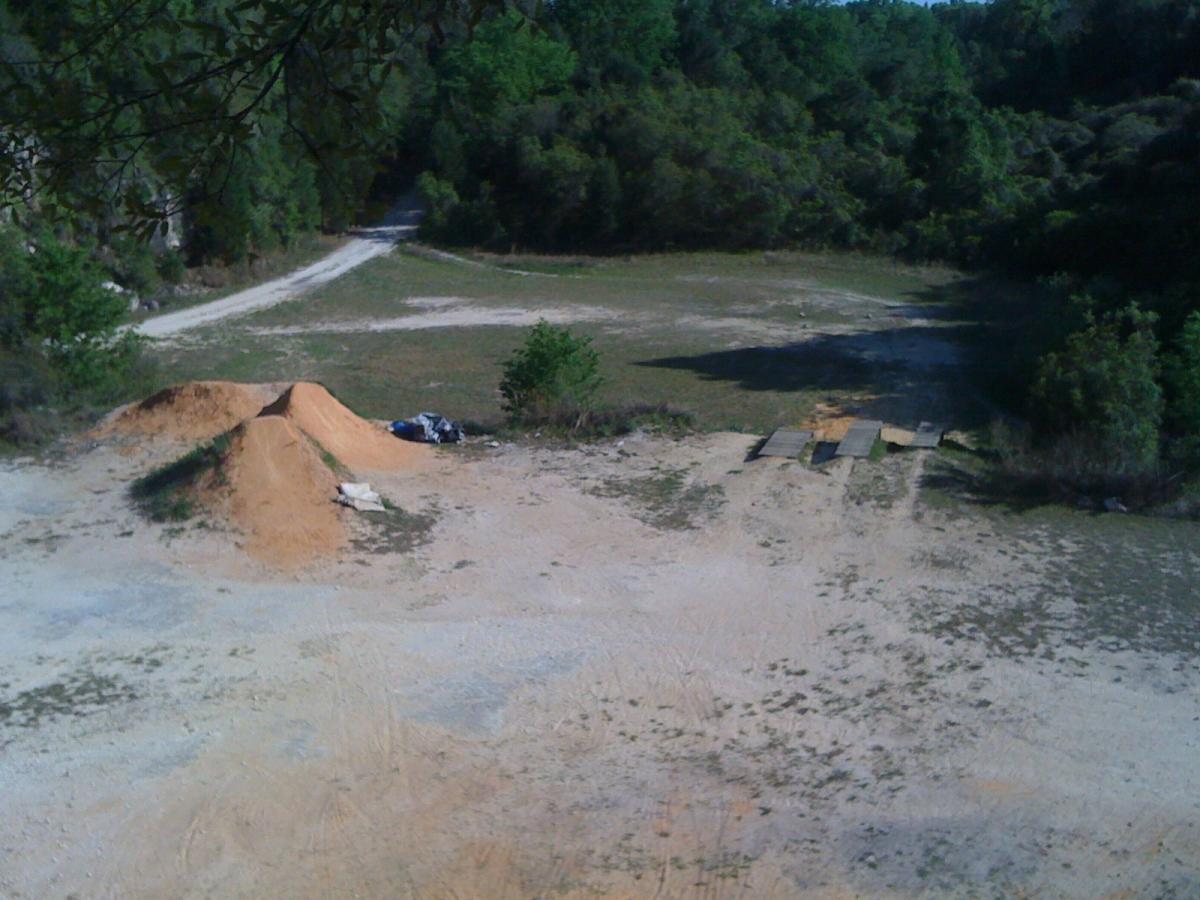 A dirt track area featuring two large dirt jumps and a set of wooden ramps, surrounded by greenery and trees in the background. A gravel path leads into the scene, and there are remnants of materials, such as tarps, scattered nearby on the ground. Santos mountain bike trail.