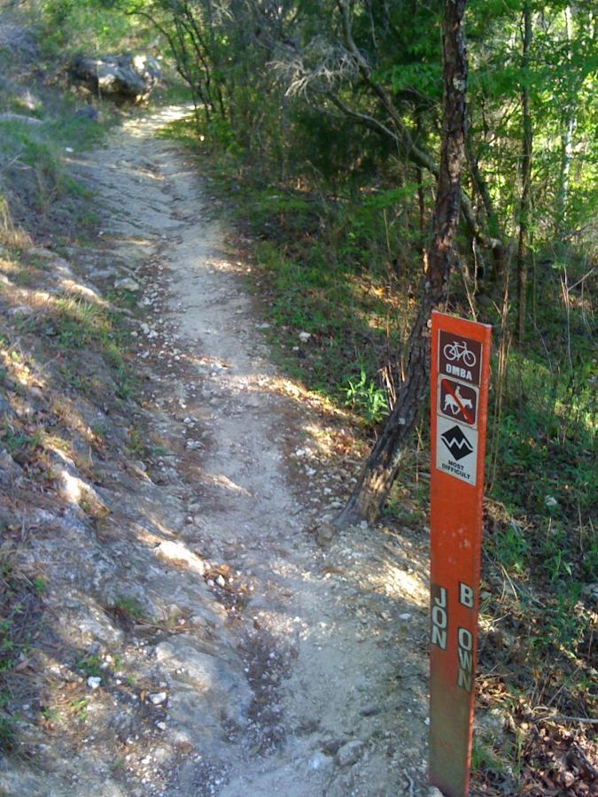A narrow dirt trail winding through a lush, green landscape, with a trail marker post on the right side. The post features symbols for biking and hiking, indicating the trail's designated uses. Santos mountain bike trail.