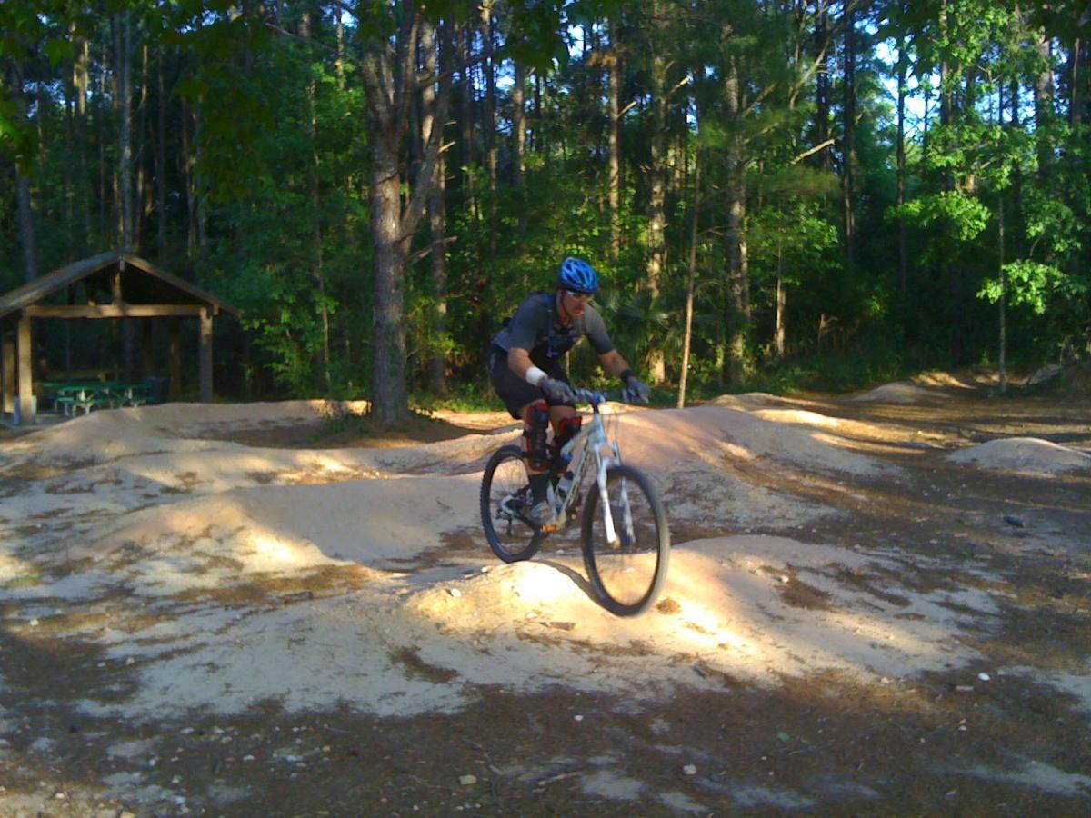 A cyclist in a helmet navigating a dirt bike trail, jumping over a small mound, surrounded by trees in a natural outdoor setting. A shelter with picnic tables can be seen in the background. Santos mountain bike trail.