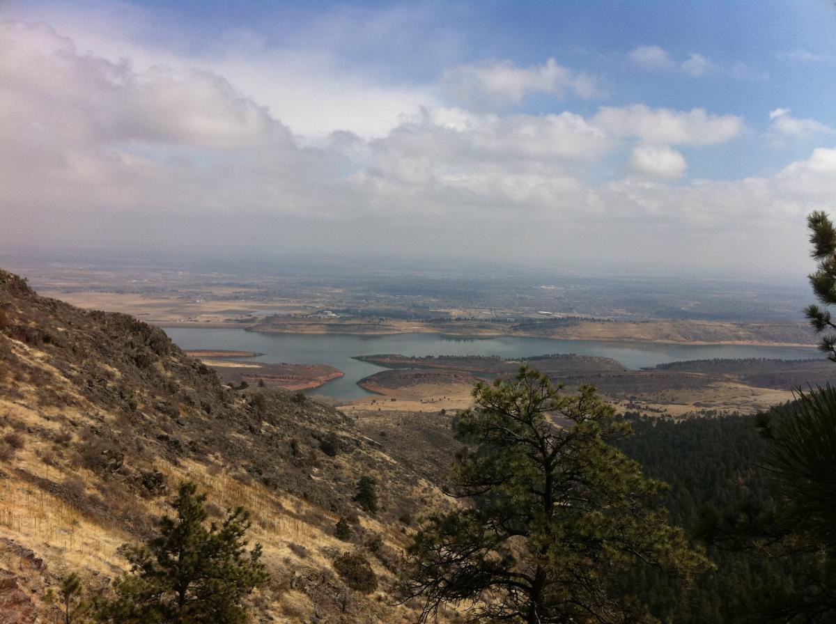 A panoramic view of a mountainous landscape featuring a river winding through the valley below, dotted with islands and grasslands. The foreground showcases rocky terrain with sparse vegetation, while the background reveals a cloudy sky and the expansive landscape stretching towards the horizon. Lory State Park mountain bike trail.