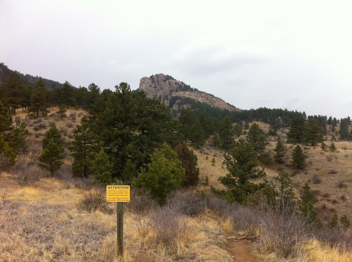 A scenic view of a hilly landscape featuring evergreen trees and dry grass under an overcast sky, with a prominent rock formation in the background. A yellow caution sign is positioned in the foreground, indicating warnings for hikers. Lory State Park mountain bike trail.