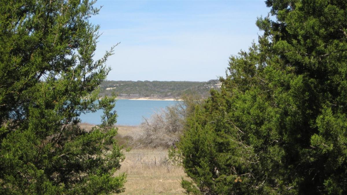 A scenic view of a lake framed by lush green trees, with rolling hills in the background under a clear blue sky. Blora mountain bike trail.