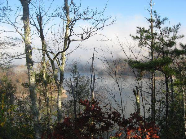 A serene view of a lake surrounded by trees, with fog gently rising over the water. The scene captures a peaceful, early morning atmosphere with bare branches and hints of autumn foliage. Tsali Thompson Loop mountain bike trail.