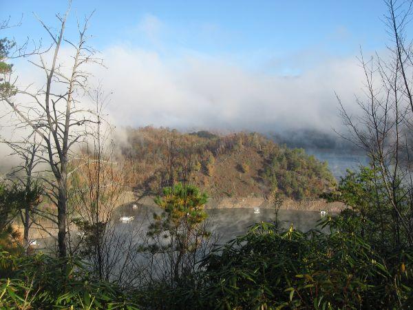 A scenic view of a misty landscape featuring a partially fog-covered mountain, with patches of greenery visible on the slopes. In the foreground, there are sparse trees and foliage, while the background reveals a dramatic sky with a mix of clouds and sunlight breaking through. The overall atmosphere is tranquil and picturesque, suggesting a serene natural environment. Tsali Thompson Loop mountain bike trail.