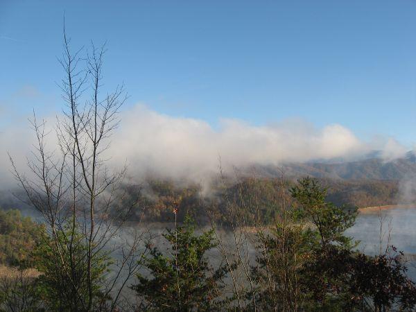 A serene landscape depicting a body of water surrounded by forested hills. The scene features low-lying clouds partially shrouding the mountains in the background under a clear blue sky. Leafless trees and greenery are visible in the foreground, creating a tranquil natural setting. Tsali Thompson Loop mountain bike trail.