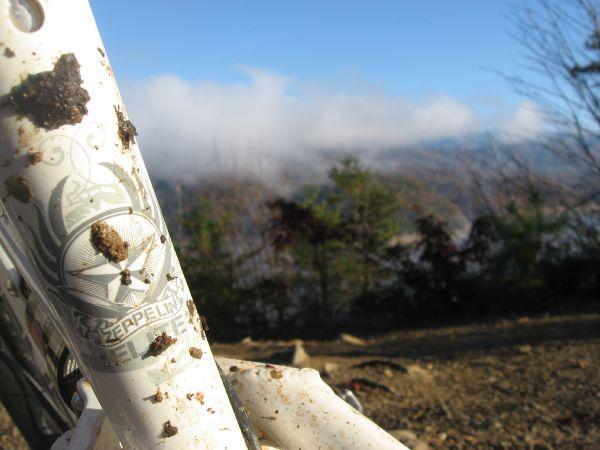 Close-up of a muddy white bicycle frame featuring a logo, with a scenic view of foggy hills and trees in the background under a blue sky. Tsali Thompson Loop mountain bike trail.