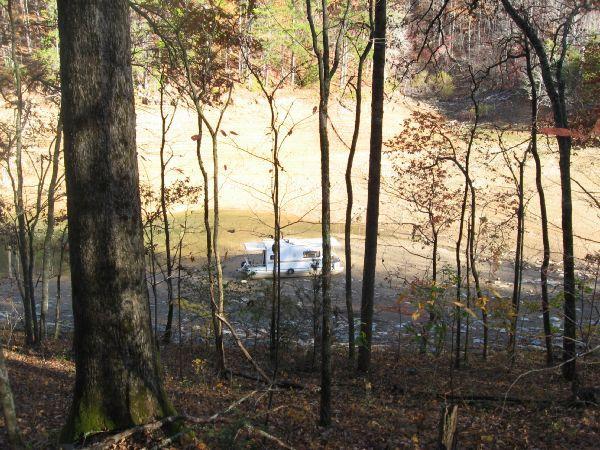 A view through trees showing a white boat in shallow water, surrounded by autumn foliage. The background features a dry lakebed and dense forest on the hillside. Tsali Thompson Loop mountain bike trail.