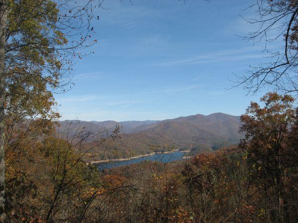 A scenic view of rolling mountains covered in autumn foliage, with a calm lake reflecting the sky. The background features a clear blue sky and distant hills, while trees with colorful leaves are visible in the foreground. Tsali Thompson Loop mountain bike trail.