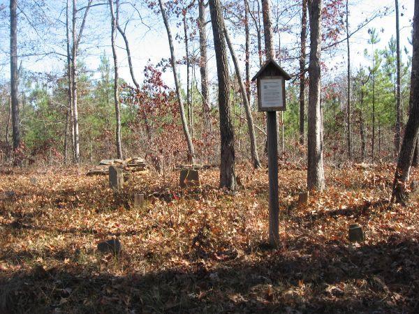A forested area with a few scattered grave markers partially covered by leaves. In the center, there is a wooden sign on a post providing information about the site. The trees in the background have sparse foliage, indicating it is likely autumn or winter. Latham Cemetary mountain bike trail.