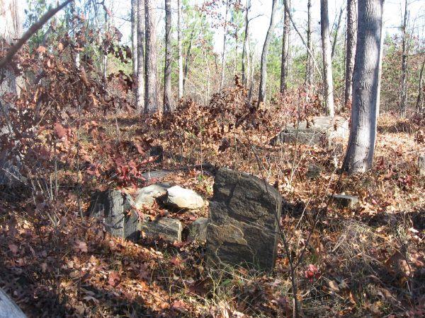 A weathered stone grave marker partially obscured by fallen leaves and surrounded by trees in a forested area, indicating an old, abandoned site. Latham Cemetary mountain bike trail.