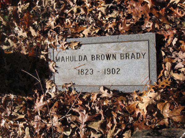 A gray gravestone with the inscription "Mahulda Brown Brady 1823 - 1902," partially covered by a layer of fallen autumn leaves. Latham Cemetary mountain bike trail.