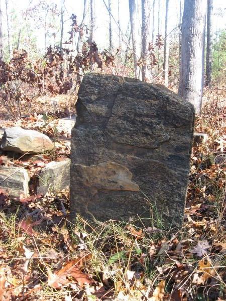 A weathered stone monument standing in a wooded area, surrounded by fallen leaves and small rocks. The stone is partially covered in moss and lichen, indicating age, with trees in the background. Latham Cemetary mountain bike trail.
