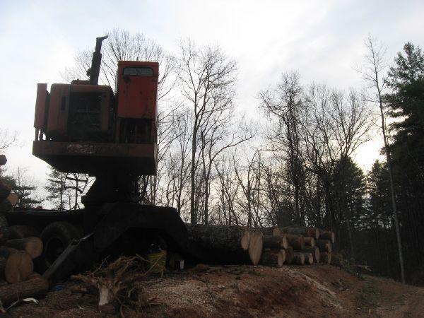 A logging machine is positioned on a hill overlooking several stacked logs, with a backdrop of bare trees against a cloudy sky. Montgomery Creek Trail mountain bike trail.