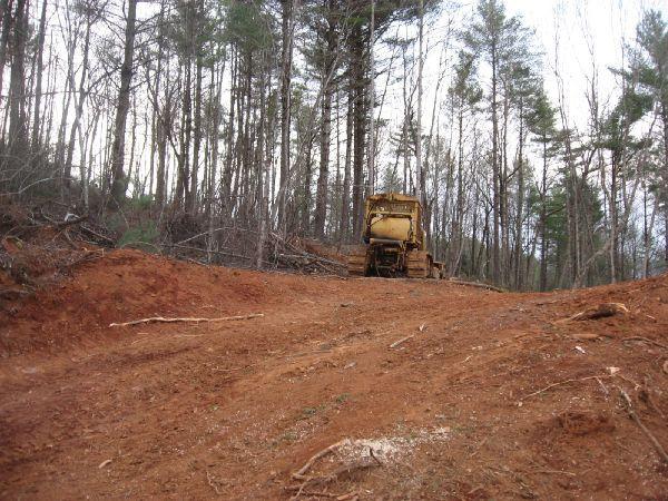 Old machinery parked on a dirt path surrounded by a forest of tall trees. The ground is reddish-brown and appears recently disturbed, with some fallen branches scattered throughout the area. Montgomery Creek Trail mountain bike trail.