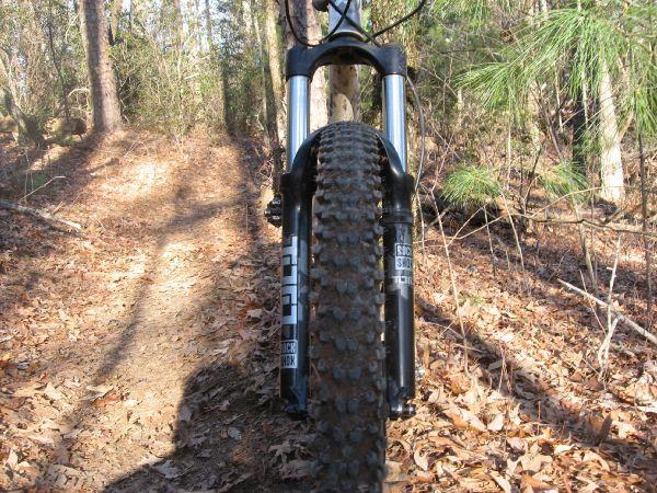 Close-up view of a mountain bike's front tire on a dirt trail surrounded by trees and fallen leaves. The trail leads into a wooded area with dappled sunlight filtering through the branches. Jake to Bull Mountain Connecter mountain bike trail.