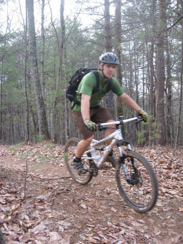 A person riding a mountain bike on a dirt trail in a wooded area, surrounded by trees and fallen leaves. The rider is wearing a helmet, a green shirt, and brown shorts, and has a backpack for carrying supplies. Black Branch Connector mountain bike trail.