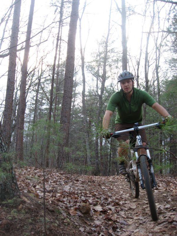 A person riding a mountain bike on a dirt trail surrounded by trees, wearing a green shirt and a helmet. The ground is covered with fallen leaves, and the scenery is lush with tall trees in the background. Black Branch Connector mountain bike trail.