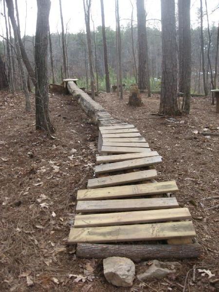 A narrow, winding wooden pathway made of planks connects two points across a forested area. The path is surrounded by tall trees and pine needles cover the ground. In the background, some greenery is visible, and several tree stumps are present along the sides of the trail. The overall ambiance is natural and rustic. Big Creek mountain bike trail.